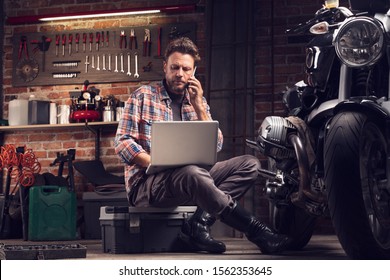 Mechanic or biker working in a workshop on a vintage motorcycle sitting talking on a mobile as he researches information on his laptop - Powered by Shutterstock