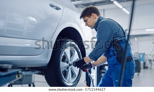 Mechanic in Blue Overalls is Unscrewing Lug Nuts
with a Pneumatic Impact Wrench. Repairman Works in a Modern Clean
Car Service. Specialists Removes the Wheel in Order to Fix a
Component on a
Vehicle.