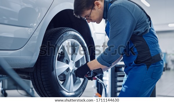 Mechanic in Blue Overalls is Unscrewing Lug Nuts\
with a Pneumatic Impact Wrench. Repairman Works in a Modern Clean\
Car Service. Specialists Removes the Wheel in Order to Fix a\
Component on a\
Vehicle.