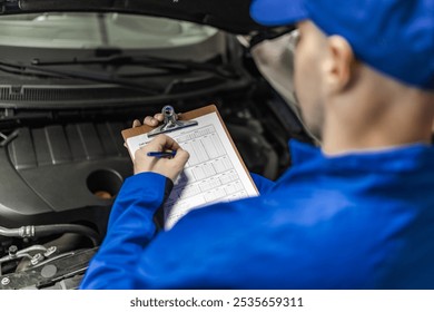 A mechanic in a blue uniform inspects a car engine while taking notes on a clipboard - Powered by Shutterstock