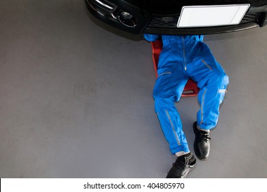Mechanic in blue uniform lying down and working under car at the garage.
