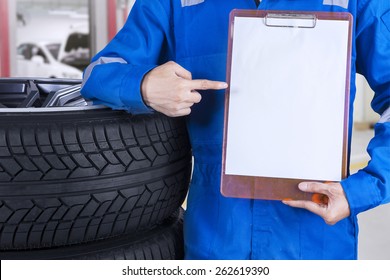 Mechanic With A Blue Work Wear Showing An Empty Copyspace While Standing Next To Tires