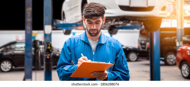 Mechanic in blue work wear uniform checks the vehicle maintenance checklist with blur lifted car in the background. Automobile repairing service, Professional occupation.