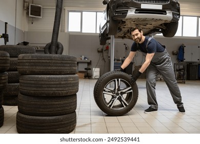 Mechanic changing tire in auto repair shop with car lifted in background. Worker in uniform and gloves holding tire surrounded by stacks of tires. Professional automotive maintenance service concept. - Powered by Shutterstock