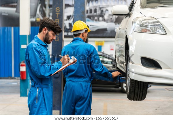 Mechanic checks the car fixing list\
while his assistant lifting the white car for examining the bottom.\
Auto car repair service center. Professional\
service.