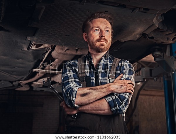 Mechanic crossed hands while standing under\
lifting car in a repair\
garage.