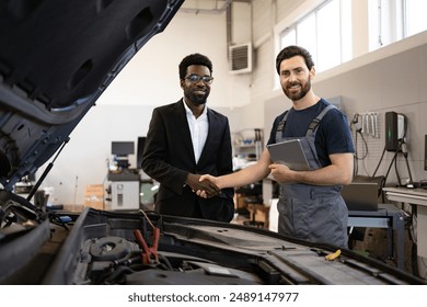 Mechanic and customer shaking hands in auto repair shop, sealing deal and expressing satisfaction. Mechanic holding tablet, customer dressed in business attire. Car hood open, tools visible. - Powered by Shutterstock