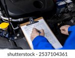 A mechanic is diligently checking car maintenance logs while examining the engine compartment of a vehicle in an auto repair shop under bright lighting.