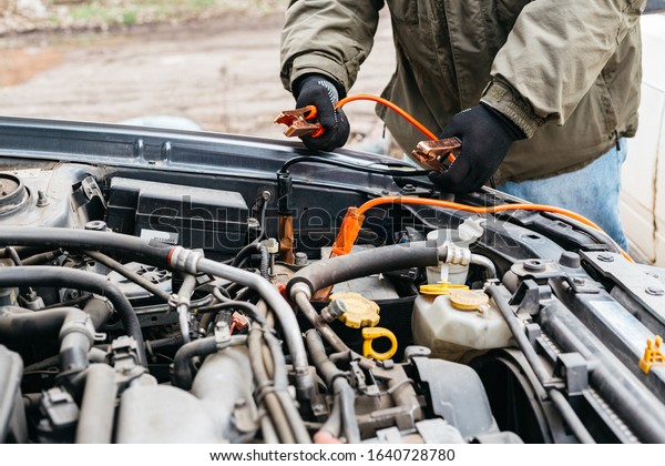 Mechanic engineer charging car battery with\
electricity using jumper cables outdoors. Red and black Jumper\
cables in male hands of car mechanic. Man in gloves working in car\
repair service\
station.