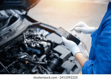 Mechanic examining car engine with mobile phone at auto repair shop. Close-up shot of male technician taking a break and texting on smartphone. Horizontal photo with workshop background. - Powered by Shutterstock