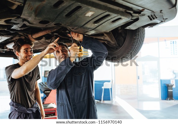 Mechanic\
fixing the car with coworker pointing and smiling. Two auto repair\
men working under a lifted vehicle in\
garage.