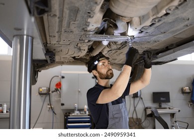 Mechanic fixing car underside with tools in auto repair shop wearing gloves and headlamp. Focus on maintenance and repair. Professional technician at work