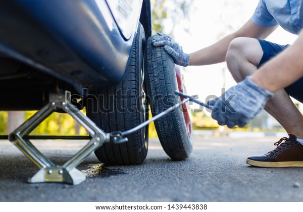 Mechanic in gloves lifts the car\
Jack on the side of the highway. Man is preparing to change the\
flat wheel lifting the car with a Jack on the side of the\
highway.