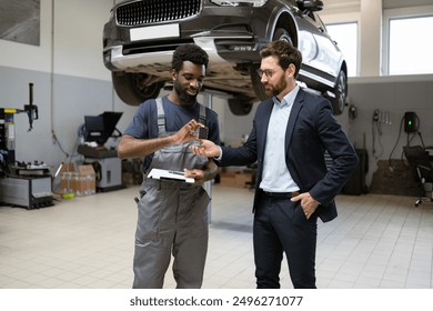 Mechanic handing car keys to businessman inside auto repair shop. Car on lift in background, discussing vehicle maintenance. Professional service, customer satisfaction, and repair expertise. - Powered by Shutterstock