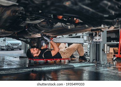 The mechanic holding wrench in hand for fix car. Strong Man fix under the car in garage with wrench. Mechanic lying down and working under the car in store. Side view.