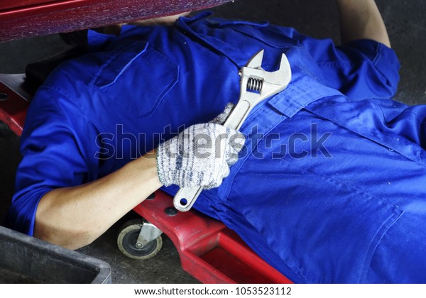 The mechanic\
holding wrench in hand for fix car. Strong Man fix under the car in\
garage with wrench. Mechanic lying down and working under the front\
of automobile in store. 