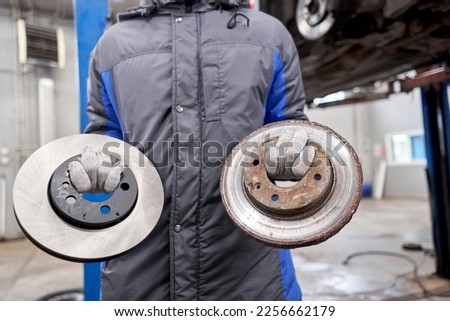 The mechanic holds old rusty brake disc and new disc. Change the old to new brake disc on car in a garage. Auto repair concept.