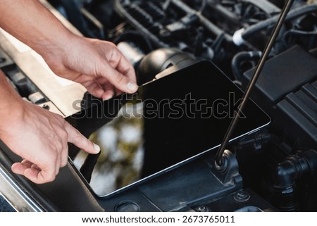 A mechanic is inspecting a car engine while utilizing a tablet to check diagnostic information. The focus is on the interaction between technology and automotive repair.