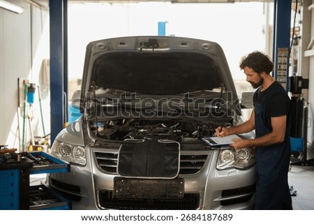 Mechanic is inspecting the engine of a car while taking notes on a clipboard in a well-equipped repair shop. Tools are organized nearby for easy access.