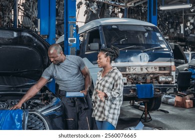 A mechanic inspects a car engine while a client observes in an automotive workshop. The space is filled with tools and vehicles, indicating a busy working environment. - Powered by Shutterstock