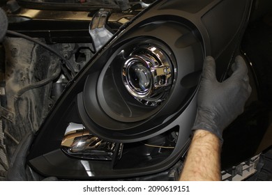 Mechanic, Installs The Headlight On The Car. Repair Of Car Headlights. Tuning And Restoration Of Automotive Optics. A Mechanic Wearing Rubber Gloves Installs An LED Lens Into The Headlight Housing.