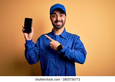 Mechanic man with beard wearing blue uniform and cap holding smartphone showing screen very happy pointing with hand and finger - Powered by Shutterstock