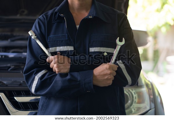 Mechanic man ready to\
wear\
Mechanic\'s set. Hold the iron Double Open End Wrench and\
Socket Wrench on both sides. Blurred image of an open car engine\
compartment.