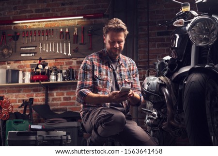 Mechanic pausing to read a message on his mobile phone as he kneels alongside a vintage motorcycle that he is repairing or servicing in a workshop