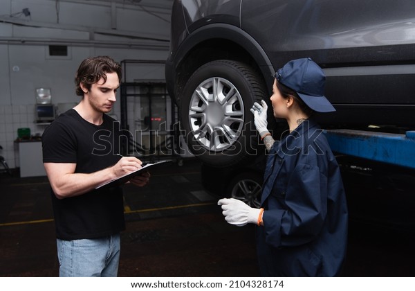 Mechanic pointing at car wheel near colleague\
writing on clipboard in\
service