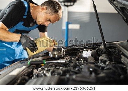 The mechanic is pouring oil into the engine. Pouring fresh oil to car engine, oil change to auto. Caucasian man in blue overalls pouring oil from plastic container.