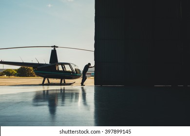 Mechanic Pushing The Door Of A Hangar With A Helicopter. Pilot Opening The Helicopter Hangar Door.