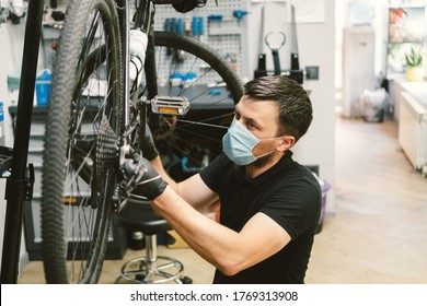 Mechanic Repairing Bicycle In Workshop Wearing Medical Mask Covid 19. Man Repair Bike On Quarantined Coronavirus Pandemic In Protective Face Mask. Man Examines And Fixes Cycle. Sport Shop Concept.