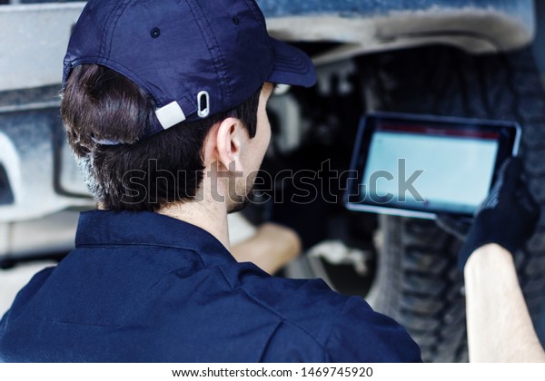 Mechanic is repairing car at service station.\
Repairman is conducting diagnostics and detecting problems by\
special software on tablet at workshop auto repair shop. Vehicle on\
hydraulic lift is\
above.
