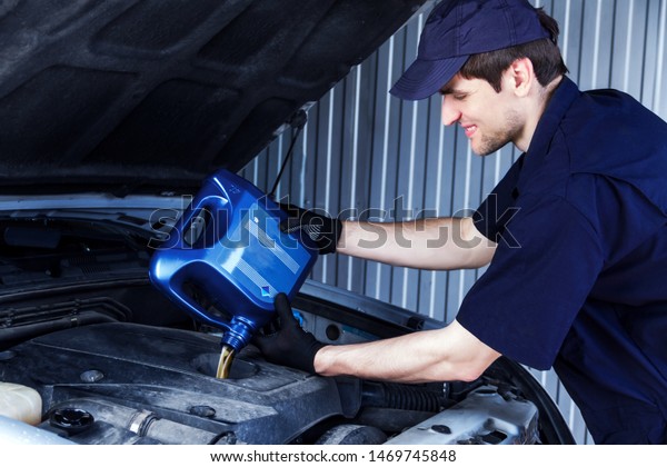 Mechanic is\
repairing, conducting diagnostics of car at service station.\
Repairer is holding blue canister and filling fueling engine oil in\
motor. Vehicle in workshop auto repair\
shop.