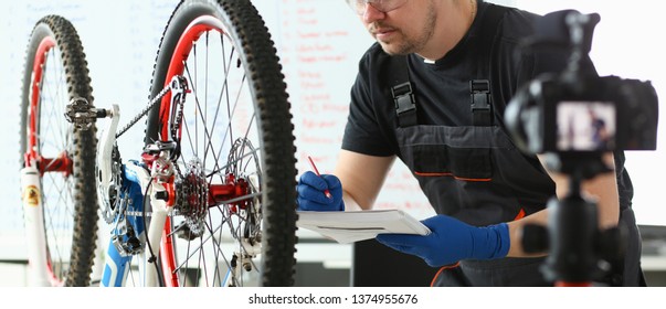 Mechanic Repairing Mountain Cycle At Sport Store. Male Blogger In Black Workwear, Protective Glasses And Gloves Fixing Bike In Repair Shop. Professional Worker Making Notes, Transport Expertise