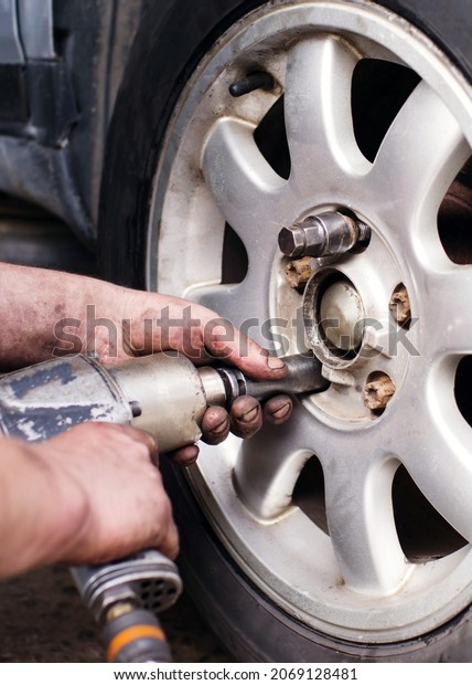 A mechanic at a\
service station replaces a car wheel, a worker\'s hands in black\
fuel oil and a tool for unscrewing bolts close-up near the wheel.\
Tire service, workflow
