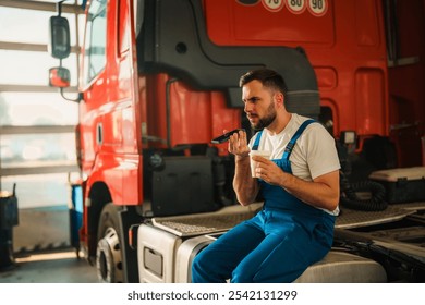 A mechanic sitting on a truck's engine, talking on a phone. He wears blue overalls. - Powered by Shutterstock