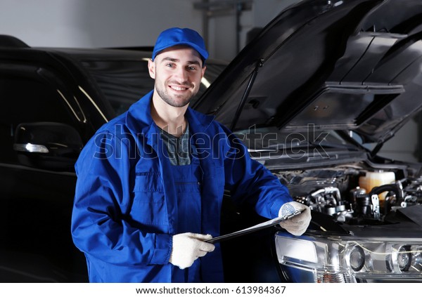 Mechanic standing in front of open car hood\
with clipboard