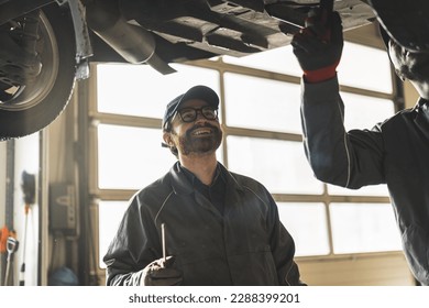 a mechanic with sunglasses looking at his colleague working underneath a car and smiling, car repair shop. High quality photo - Powered by Shutterstock