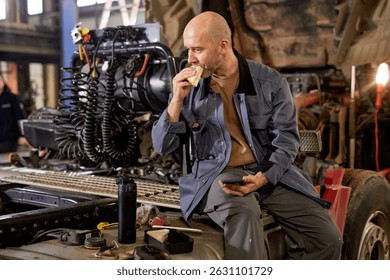 Mechanic taking a break while sitting on a vehicle with engine components visible behind. Eating a snack and holding a smartphone, surrounded by auto tools and equipment in workshop