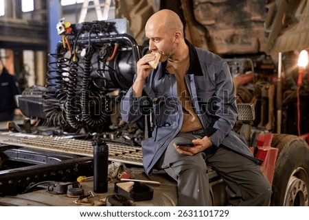 Mechanic taking a break while sitting on a vehicle with engine components visible behind. Eating a snack and holding a smartphone, surrounded by auto tools and equipment in workshop