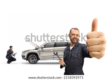 Mechanic in a uniform holding a wrench and gesturing thumbs up and other worker checking a SUV in the back isolated on white background