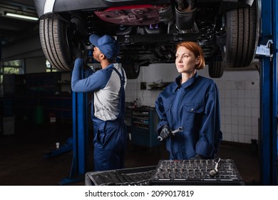 Mechanic in uniform holding wrench while african american colleague working with car in garage