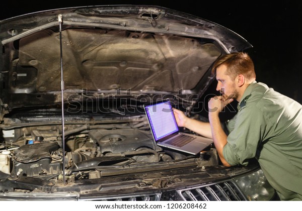 Mechanic in uniform looking intently at the\
laptop is connected to the motor vehicle for testing and\
adjustment. Mechanic carries out maintenance of the car. The\
removal of bugs of the\
engine.