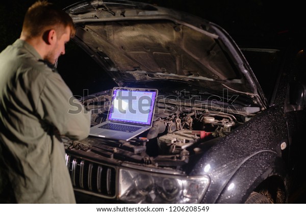Mechanic in uniform looking intently at the\
laptop is connected to the motor vehicle for testing and\
adjustment. Mechanic carries out maintenance of the car. The\
removal of bugs of the\
engine.