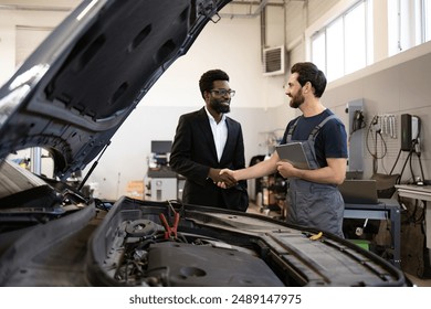 Mechanic in uniform shaking hands with customer in suit near car engine in auto shop. Concept of service, professionalism, satisfaction, and business relationship in automotive industry. - Powered by Shutterstock