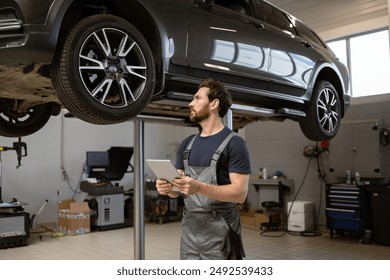 Mechanic in uniform using tablet while inspecting car on lift in garage. Professional workshop setting with tools and equipment visible. - Powered by Shutterstock