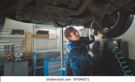 Mechanic unscrewing parts of automobile's bottom under lifted car