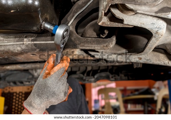 A mechanic unscrews an oil drain plug\
underneath the chassis of a SUV with a ratchet wrench. First step\
of an oil change. Maintenance\
procedure.