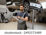 Mechanic using digital tablet in car repair shop with raised vehicle in background. Man in work uniform checking diagnostics. Auto service, modern technology in automobile maintenance.